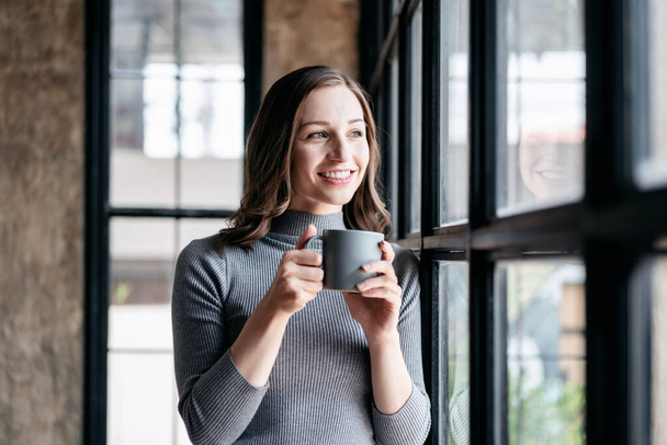 Bizneswoman przytulny styl życia koncepcja, Businesswoman picia gorącej czekolady i stoi w pobliżu okna do patrząc na zewnątrz zrobić przerwę z korzystaniem podczas relaksu po pracy. - Zdjęcie, obraz
