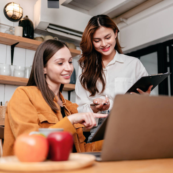 Twee jonge vrouw lezen van zakelijke informatie in klembord en digitale tablet samen om te brainstormen over nieuwe project business terwijl het werken met laptop op de tafel in de keuken thuis. - Foto, afbeelding