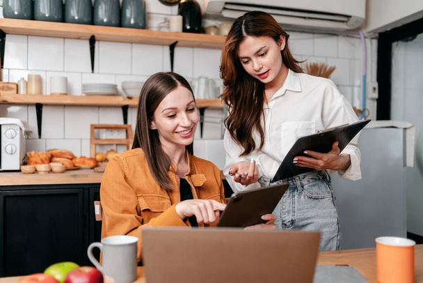 Zwei junge Frauen lesen gemeinsam Geschäftsinformationen in Klemmbrett und digitalem Tablet, um über neue Projektgeschäfte nachzudenken, während sie zu Hause mit Laptop auf dem Tisch in der Küche arbeiten. - Foto, Bild