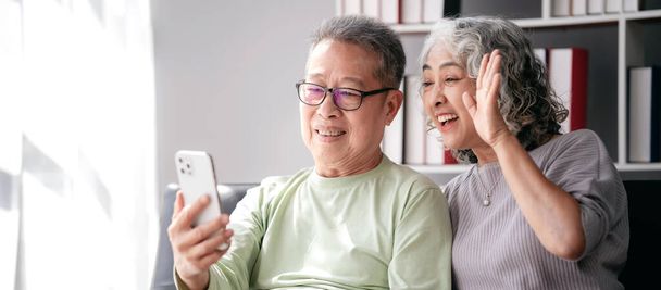 Seniorenpaar nutzt Smartphone zur Videokonferenz und zur Begrüßung mit der Familie, während es auf der Couch sitzt und Zeit für Aktivitäten zur Entspannung im Lebensruhestand zusammen im heimischen Wohnzimmer verbringt. - Foto, Bild