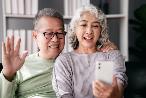Vanhempi pari käyttää älypuhelinta videoneuvotteluun ja tervehtimiseen perheen kanssa istuen sohvalla ja viettäen aikaa rentoutumiseen elämässä eläkkeelle yhdessä kodin olohuoneessa. - Valokuva, kuva
