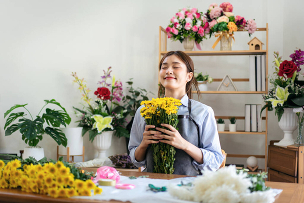 Γυναίκα ανθοπώλης με ποδιά που κρατά κίτρινο χρυσάνθεμα και μυρίζει το λουλούδι με φρεσκάδα ενώ δημιουργεί και σχεδιάζει λουλουδάτο μπουκέτο λουλουδιών σε σύνθεση στο ανθοπωλείο της για παράδοση στον πελάτη. - Φωτογραφία, εικόνα