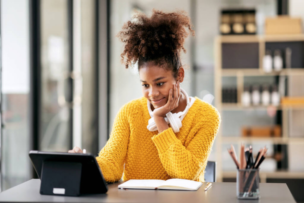 Studentessa afroamericana in maglione che guarda l'e-learning per studiare lezione online su tablet e prendere appunti mentre è seduta sulla scrivania per imparare conoscenza ed educazione all'università. - Foto, immagini
