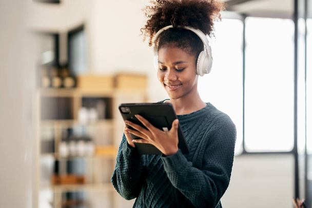 African american student woman wearing sweater and headphones to studying lesson online class and writing information on digital tablet while learning education and knowledge in university. - Фото, зображення