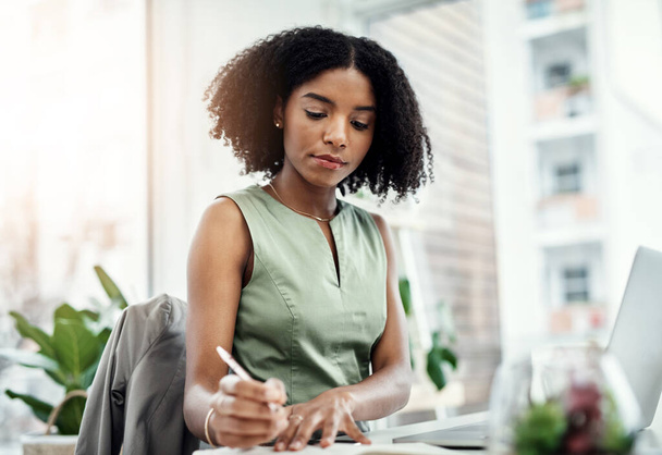 Корпоративная, черная деловая женщина пишет в книге и за ноутбуком сидит за столом в своем офисе. Планирование графика, напоминание и ориентированные африканские женщины-работницы на рабочем месте пишут заметки о информации. - Фото, изображение