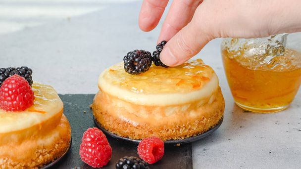 Mini kaastaart close-up. Topping cheesecakes met sinaasappelmarmelade, verse bessen, en rozemarijn, close-up versieren proces - Foto, afbeelding