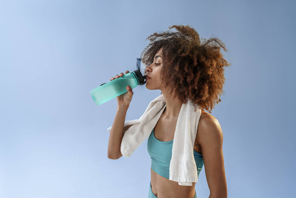 vrouw staan met water fles en handdoek rond haar nek na sportschool over studio achtergrond  - Foto, afbeelding