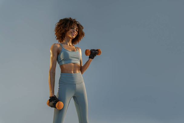 Glimlachende gespierde vrouw doen oefening met gewicht halters op studio achtergrond  - Foto, afbeelding