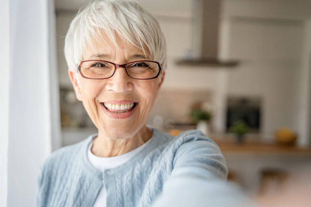 Közelkép portré egy idős nő rövid haj boldog mosoly pozitív érzelem másolat tér áll otthon beltéri szürke fehér haj önarckép szelfi vagy videohívás - Fotó, kép
