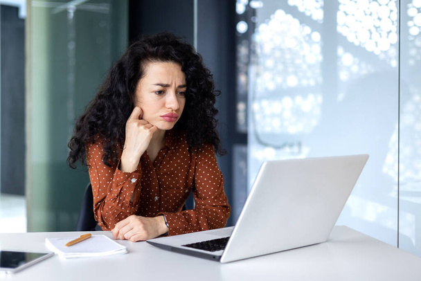 Serious thinking Hispanic businesswoman working inside modern office, female programmer looking at laptop testing new software. - Foto, Imagen