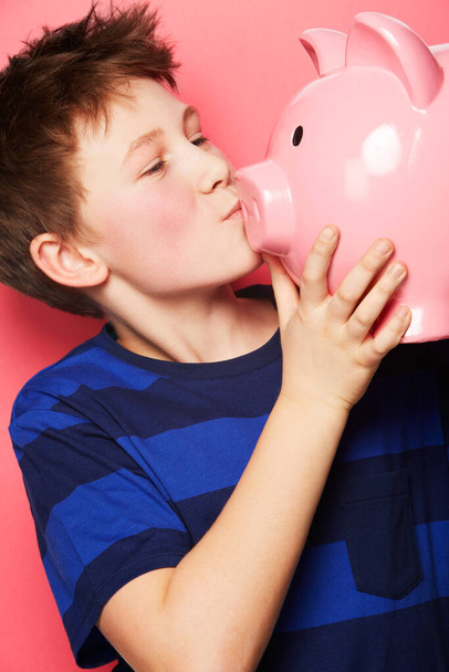 Szczęśliwy chłopiec, oszczędności i pocałunek na skarbonkę dla inwestycji, pieniędzy lub monet na różowym tle. Małe dziecko lub dziecko trzymające i całujące skarbonkę dla wolności finansowej, gotówki lub wzrostu zysku. - Zdjęcie, obraz