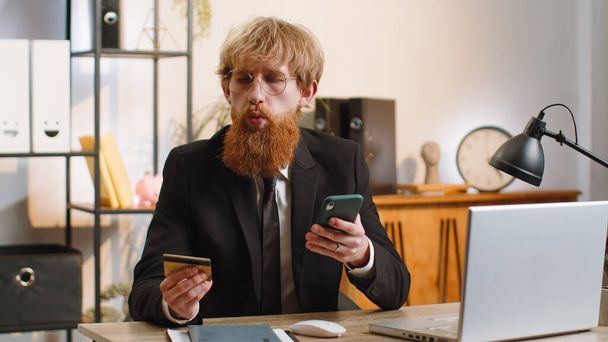 Bearded businessman programmer software developer working making online purchase payment shopping with credit bank card smartphone at modern home office desk with laptop. Young freelancer business man - Fotó, kép