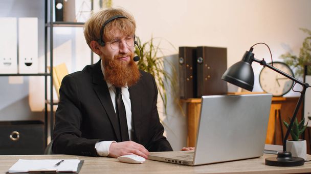 Young business man working on laptop wearing headset, freelance worker call center or support service operator helpline, having pleasant talk with client or colleague communication support at office - Фото, зображення