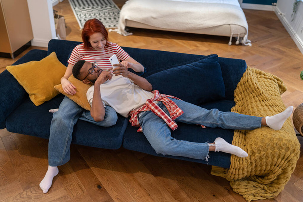 リラックスしたアフリカ系アメリカ人の男性は、リビングルームのソファで膝ヨーロッパのガールフレンドの携帯電話に横たわっています。笑顔女性休んでいます上のソファタッチスクリーンサーフィンウェブ上のスマートフォンと一緒にボーイフレンド - 写真・画像