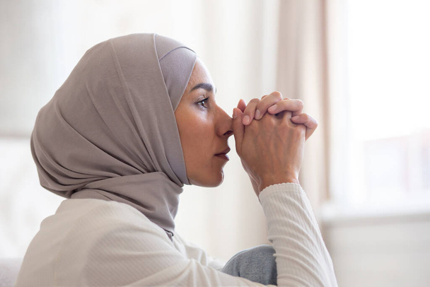 Close-up photo. Portrait of young Muslim woman in hijab looking serious and sad. He sits thoughtfully at home, folding his hands together in front of him. - Foto, imagen