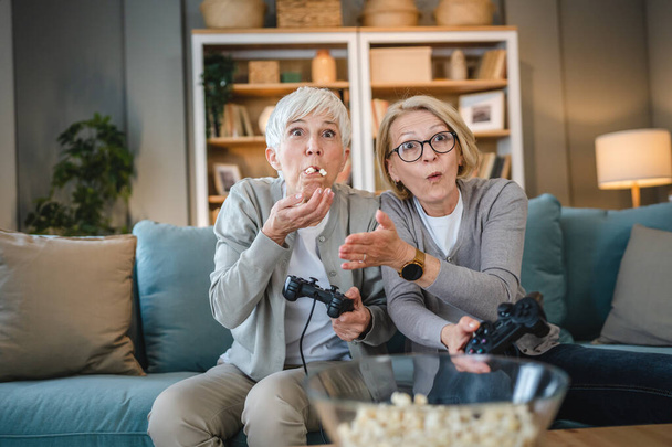 Kaksi vanhempi naisten valkoihoinen ystäviä tai sisaria onnellinen vanha sisarukset eläkeläinen pelaa videopeli konsoli ohjaimella tai ohjaimet istuessaan kotona oikeita ihmisiä perheen vapaa-ajan käsite kopioi tilaa - Valokuva, kuva