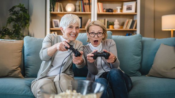 Two senior women caucasian friends or sisters happy old siblings pensioner playing video game console using joystick or controllers while sitting at home real people family leisure concept copy space - Photo, Image