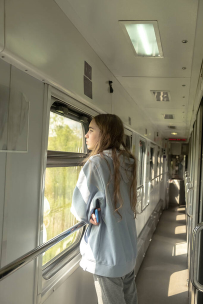 Ξανθιά έφηβη στέκεται στο διάδρομο του τρένου και κοιτάζει έξω από το παράθυρο. Σχέδιο ταξιδιού με τρένο. Κάθετη φωτογραφία  - Φωτογραφία, εικόνα