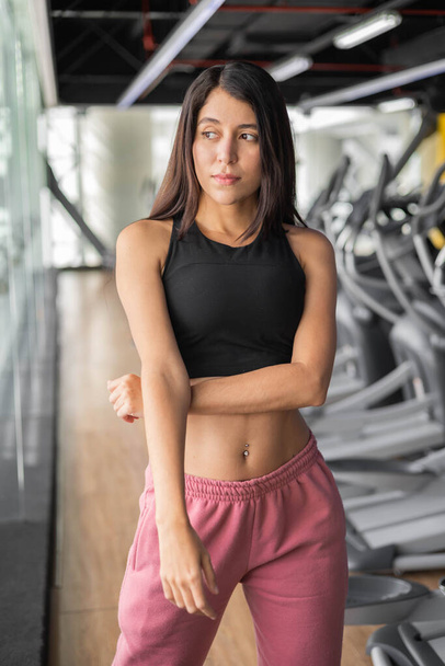 Slim female model with straight hair wears a sports top and pants, young latin woman, gym interior, healthy lifestyle and person portrait - Foto, imagen