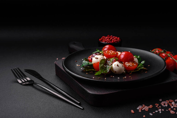 Delicious fresh salad with mozzarella cheese, cherry tomatoes, herbs, salt and spices on a dark concrete background - Photo, Image