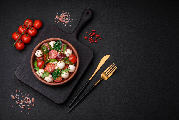 Delicious fresh salad with mozzarella cheese, cherry tomatoes, herbs, salt and spices on a dark concrete background - Photo, Image