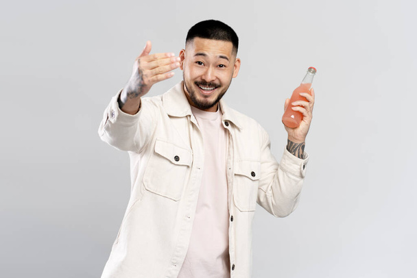 Ystävällinen hymyilevä aasialainen mies päällään rento vaatteita, tilalla pullo juomalla kutsua juhliin, eristetty valkoisella taustalla. Positiivisen elämäntavan käsite - Valokuva, kuva