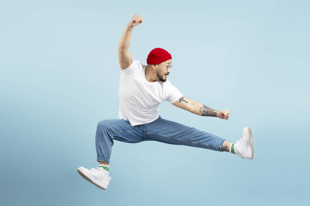 Handsome asian man, fighter with stylish tattoo, training isolated on blue background. Korean dancer wearing red hat, shoes, white t shirt, jumping high looking away. Sport, advertisement concept - Photo, Image