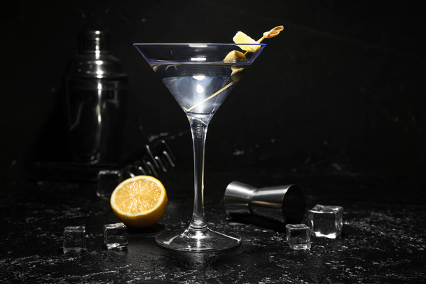 Glass of tasty martini, ice cubes, lemon and measure cup on dark background - Photo, Image