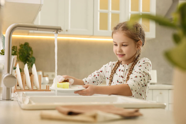 Little girl washing plate above sink in kitchen - Zdjęcie, obraz