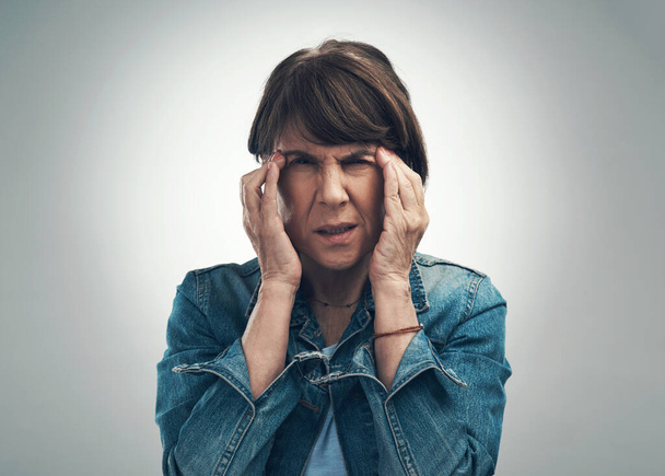 My heads spinning round and round. Studio portrait of a senior woman experiencing a headache against a grey background - 写真・画像