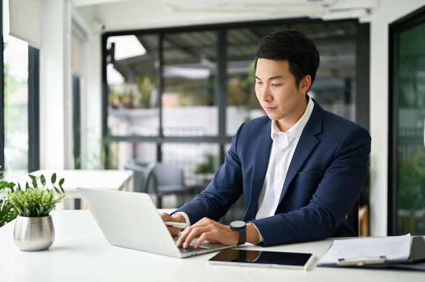 Концентрированный и умный азиатский бизнесмен тысячелетия или мужчина исполнительный директор, использующий ноутбук, управляющий своими бизнес-задачами на компьютере за своим столом. - Фото, изображение