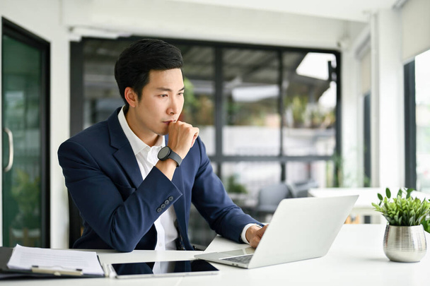 Zamyšlený a soustředěný tisíciletý asijský podnikatel nebo šéf mužského pohlaví přemýšlí o plánu, jak vyřešit problémy svého projektu, podívat se na obrazovku notebooku a pracovat ve své kanceláři. - Fotografie, Obrázek