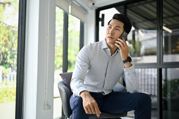 Profesjonalny milenijny azjatycki biznesmen lub męski szef rozmawia przez telefon ze swoim dostawcą biznesowym podczas siedzenia w biurze. - Zdjęcie, obraz