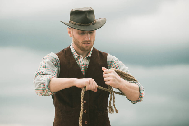 Cowboy wearing hat. Western life. Handsome bearded west farmer. Portrait of man cowboy or farmer. Western. Cowboy with lasso rope on sky background - Fotografie, Obrázek