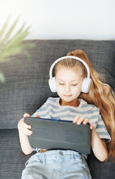 Holčička ležící na gauči s elektronickým tabletem poslouchá hudbu v bílých sluchátkách. Použití bezdrátových zařízení pro vzdělávání. - Fotografie, Obrázek