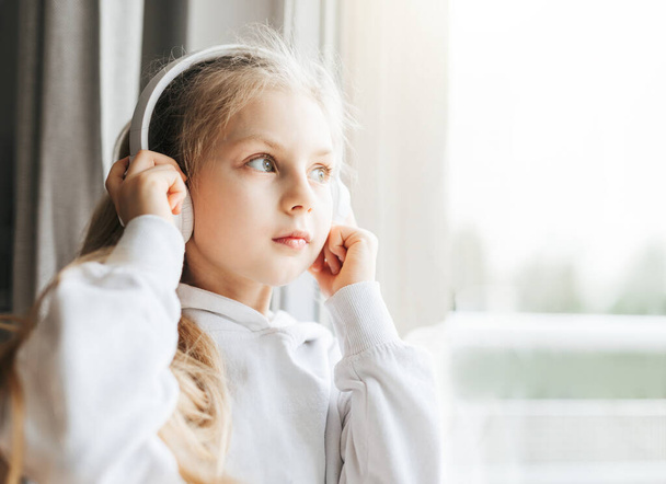 Petite fille avec écouteurs à l'intérieur à la maison, assise sur le rebord de la fenêtre et écoutant de la musique - Photo, image