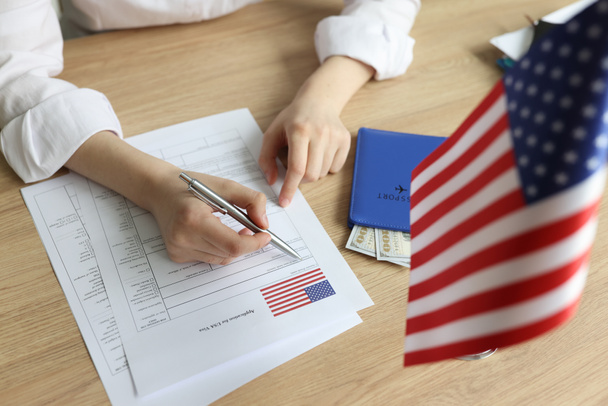 Mladá žena vyplní formulář žádosti o vízum. Ženy podepisují dokumenty, které sedí u dřevěného stolu s vlajkou USA a pasem s dolarovými bankovkami - Fotografie, Obrázek
