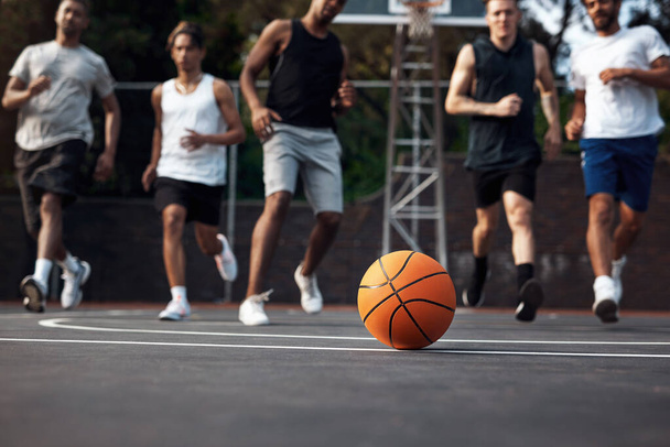 Legendy si spolu hrají. Detailní záběr basketbalu na sportovním hřišti se skupinou mladých mužů v pozadí - Fotografie, Obrázek