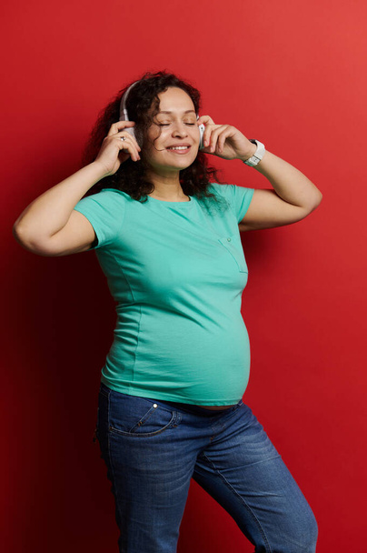 Portrait of a middle aged curly haired multi ethnic beautiful pregnant woman in turquoise t-shirt and blue jeans, enjoying soothing music, wearing wireless headphones, isolated on red background - Фото, изображение