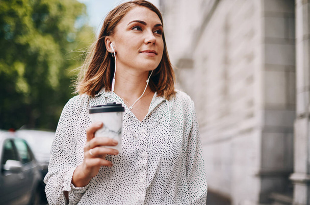 Dit gaat mijn kant op. een zakenvrouw die een koffie vasthoudt en naar muziek luistert via een oortelefoon terwijl ze door de stad loopt - Foto, afbeelding