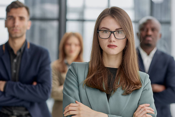 businesswoman standing with her arms crossed in an office while her colleagues have a meeting in the background. - Foto, Bild
