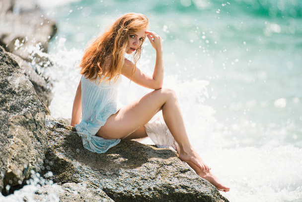 Summer dream. Sexy woman sitting on rocky beach against sea blue waves. Sensual young woman wearing sexy summer dress on a beach with her elbow on her knee and her hand to her head - Valokuva, kuva