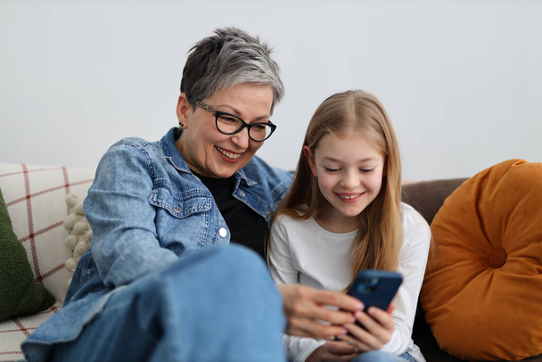 Happy grandmother and granddaughter watching fun content on phone in home interior. - Foto, Bild