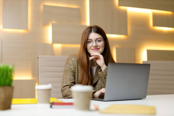 Positive woman in the workplace, a young employee sits at a table with a laptop. - Φωτογραφία, εικόνα