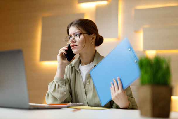A woman in the office is talking on the phone with a folder in her hands at the workplace, discussing the details of the contract. - Foto, immagini