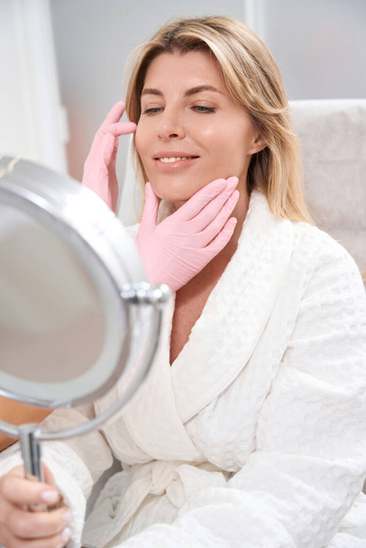 Smiling woman in a bathrobe looks in the mirror, she is at a consultation in the clinic of aesthetic medicine - Foto, imagen
