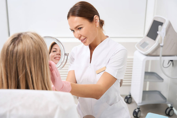 Cosmetologist in the clinic of aesthetic medicine consults a patient in a bathrobe, an employee in a medical uniform - Photo, Image
