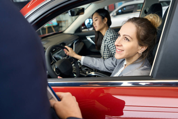 Due donne gioiose sedute sui sedili anteriori in auto rossa guardano avanti - Foto, immagini