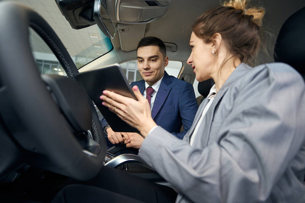 Мужчина и женщина в деловой одежде сидят на переднем сиденье в автомобиле и смотрят на планшет - Фото, изображение