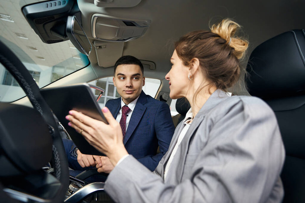 Feliz hombre y mujer en chaquetas con la tableta sentado en los asientos delanteros en el automóvil y mirándose el uno al otro - Foto, imagen